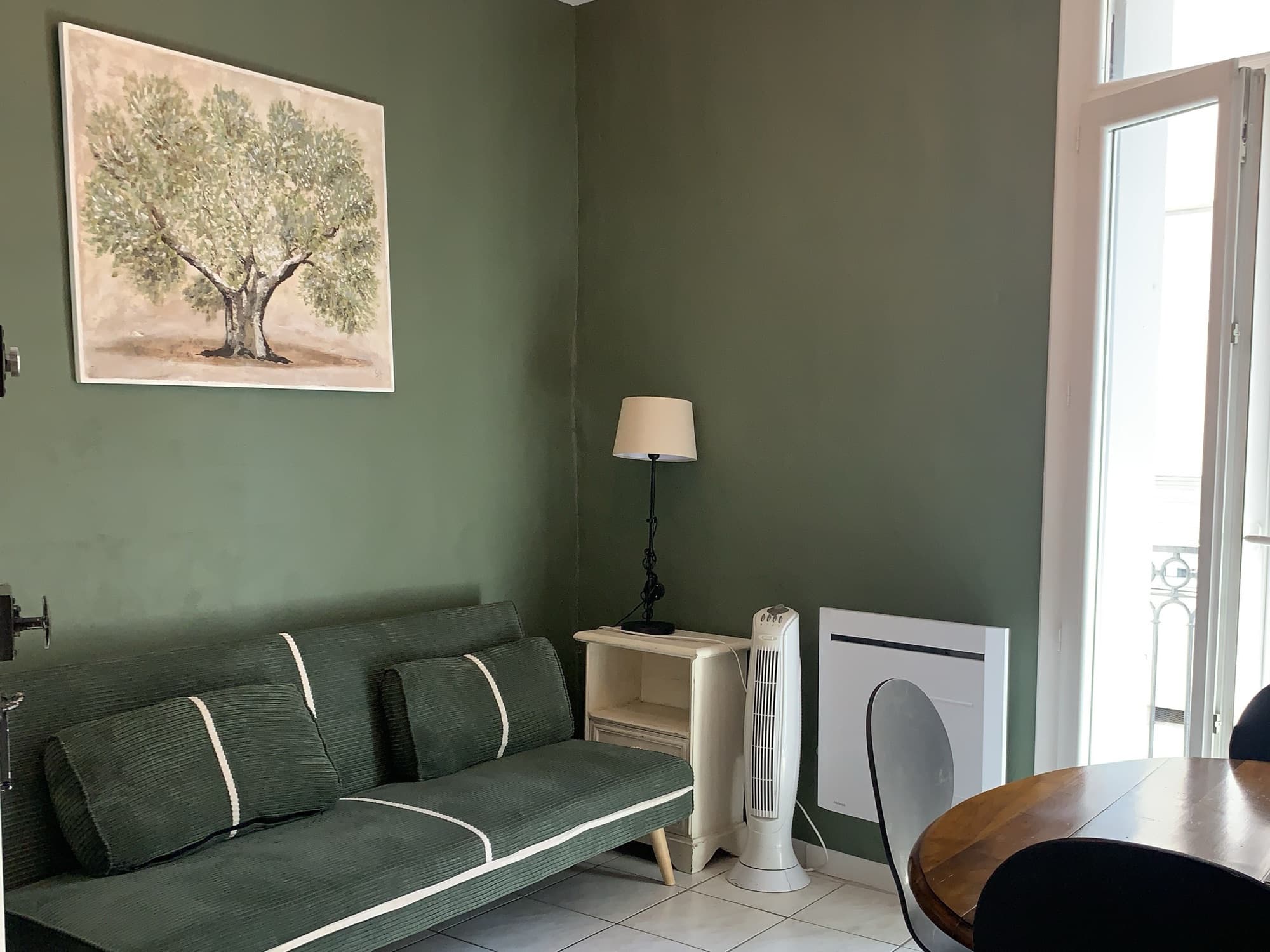 Séjour du studio rénové et meublé : canapé vert, mur vert sauge, kitchenette et coin repas devant la fenêtre